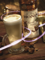 A glass of latte featuring elaborate foam art crafted by a skilled barista is placed beside a bottle of Monin Roasted Hazelnut Syrup x 1 litre. Scattered hazelnuts and a brass scoop adorn the wooden café table, enhanced by a swirling light effect that adds dynamic flair to the scene.