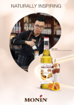 In a café bar setting, a barista adorned with glasses and an apron skillfully pours a drink. Nearby, the Monin logo is prominently featured alongside a bottle of Monin Honeycomb Syrup x 1 litre and an artfully decorated beverage. The phrase "Naturally Inspiring" accompanies the Monin branding.