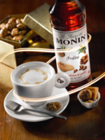 A meticulously crafted cup of coffee with foam rests on a saucer, accompanied by a spoon and praline candy. Behind it, a bottle of Monin Praline Syrup x 1litre and a box of pralines are elegantly arranged on the wooden café surface with an artistic light swirl effect.