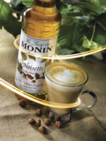 A 1-liter bottle of Monin Hazelnut Syrup is placed beside a glass mug of creamy, barista-style coffee with a frothy top. Hazelnuts are scattered on a burlap cloth backdrop, while a swirl of light creates a warm and inviting café atmosphere.