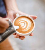 A skilled barista elegantly holds a white cup crafted from Lavazza Super Crema Coffee Beans 1kg, adorned with heart-shaped latte art. As milk cascades from a silver pitcher into the cup, the bustling café's background fades softly, highlighting the artistry and precision of the craft.