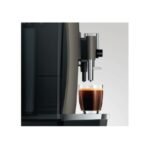 The Jura W8 - Tanked Bean to Cup Coffee Machine, with its elegant design, reminiscent of a barista's touch, dispenses coffee into a transparent mug on its silver drip tray. The softly lit background accentuates the rich, dark hue of the freshly brewed café-quality coffee.