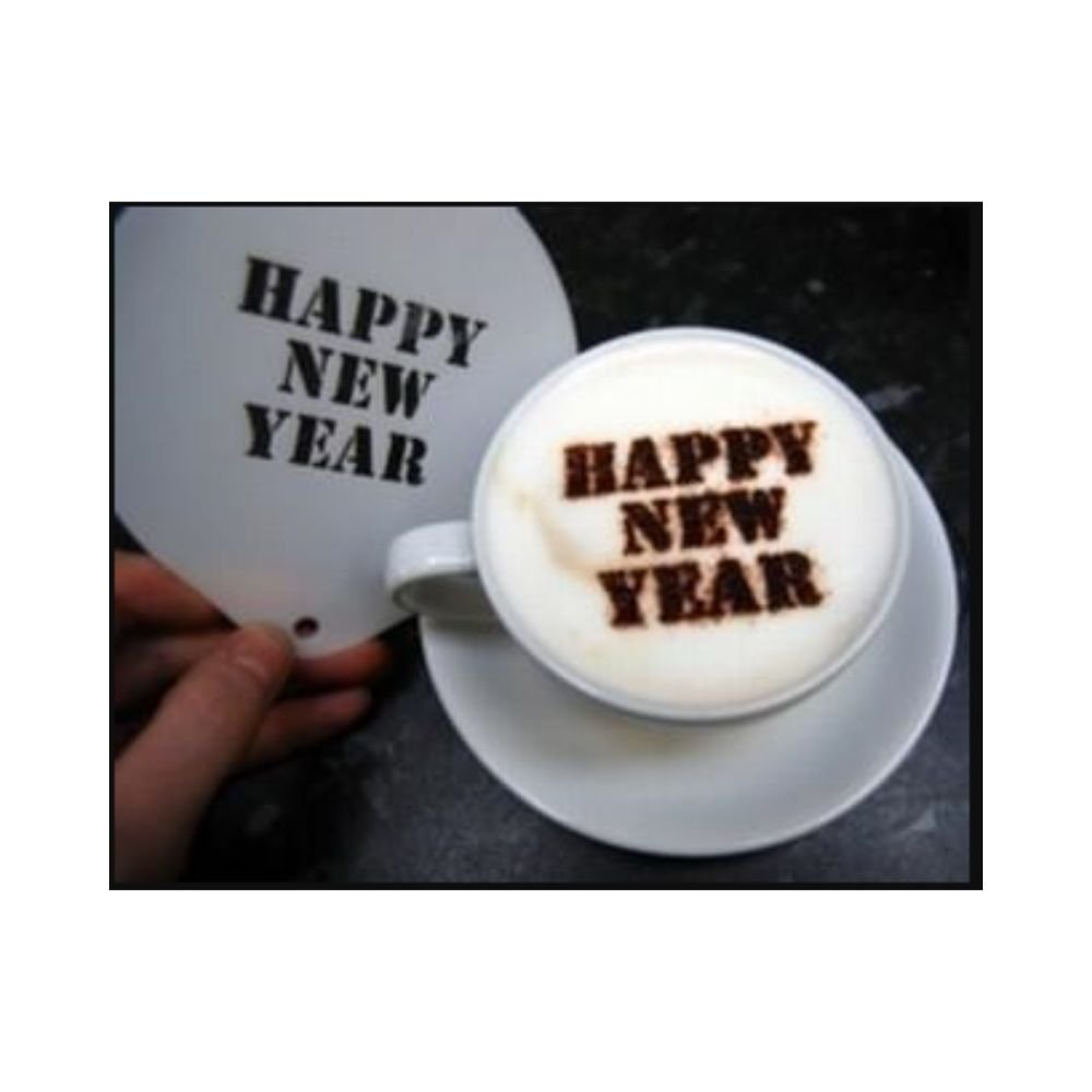 A skilled barista crafts a cappuccino with "HAPPY NEW YEAR" artfully written in cocoa on the foam, using the Stencil Happy New Year! A hand holds the stencil beside the cup, enhancing the cozy café ambiance set against a dark background.