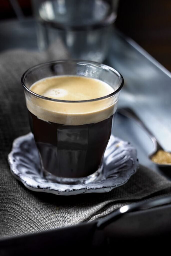 Image shows a small glass with dark and robust Americano coffee with sturdy crema on top, the glass sits on a small shell like saucer