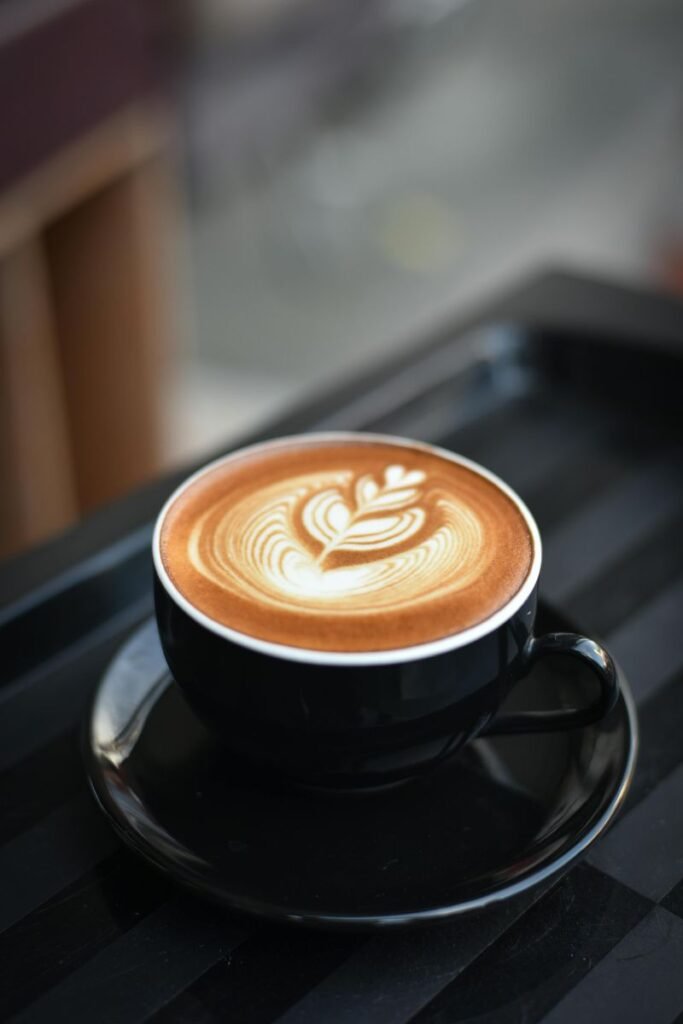 Image shows a black shiny cup and saucer with a creamy Cappuccino with leaf pattern in the froth