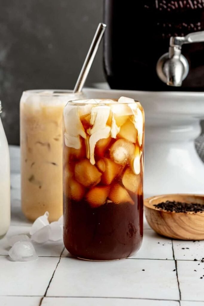 A tall can shaped glass with cold brew coffee filled with ice cubes and a drizzle of cream on a kitchen counter top