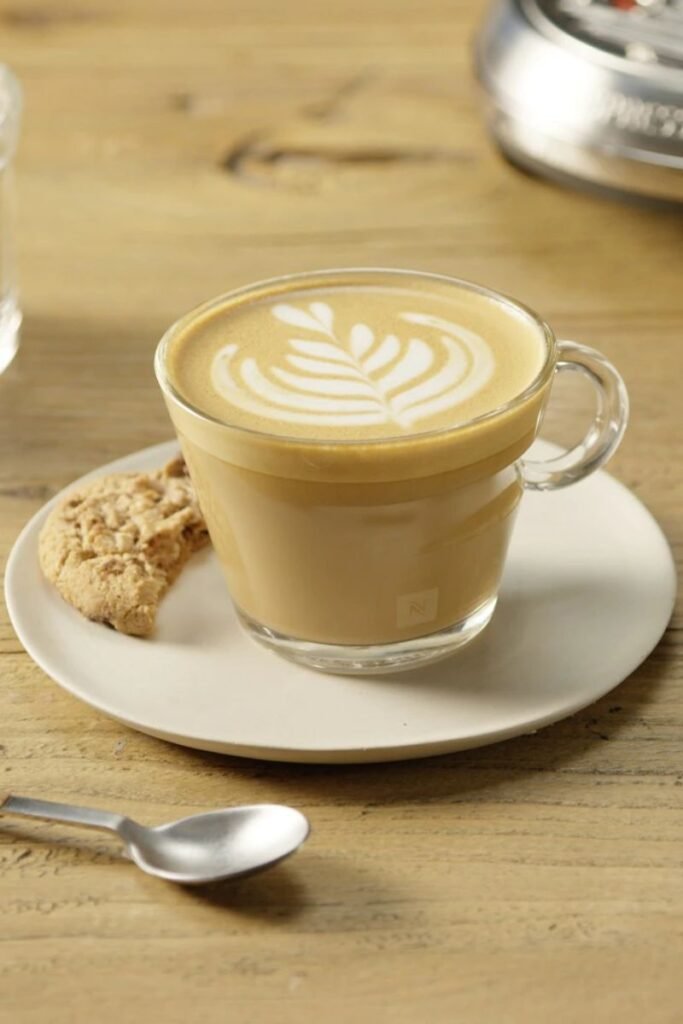 A small glass teacup with flat white coffee, creamy crema and small half eaten cookie on the white porcelain saucer