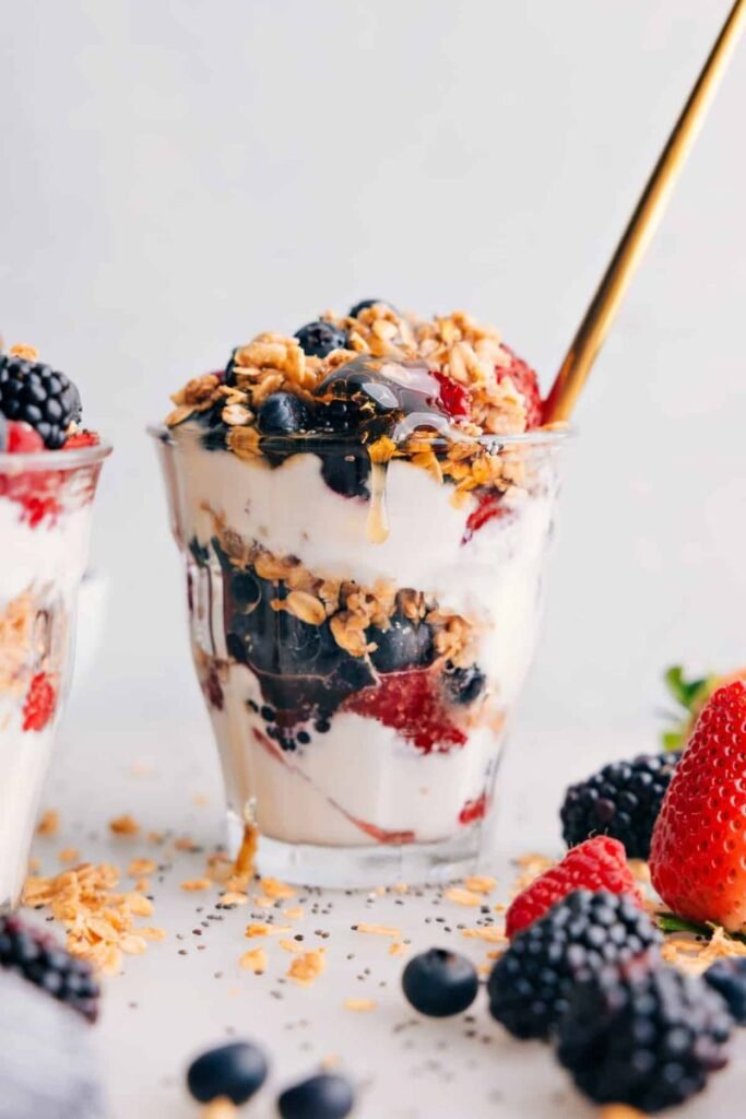 A glass with layers of Greek yoghurt, fresh fruit and granola surrounded by blackberries, blackcurrrants, and strawberries