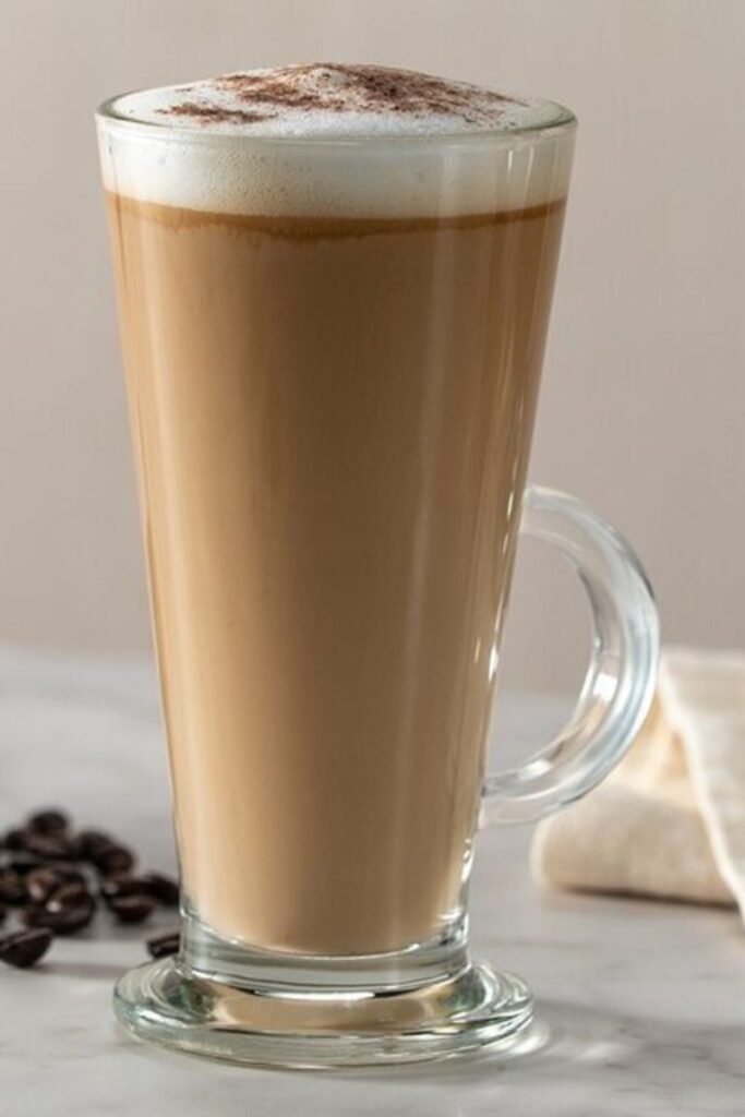 A tall glass Latte mug with a frothy head and sprinkled chocolate on top, sitting on a marble worktop with scattered coffee beans